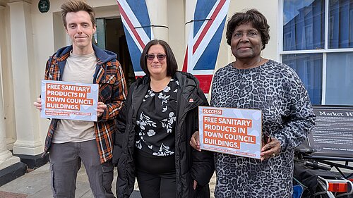 Cllr Nathan Hunt, Cllr Jo Harvey, and Cllr Padrica Pennington holding signs that read "Free Sanitary Products in Town Council Buildings"