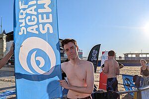 Cllr Nathan Hunt stood on a beach by a flag reading "Surfers Against Sewage"